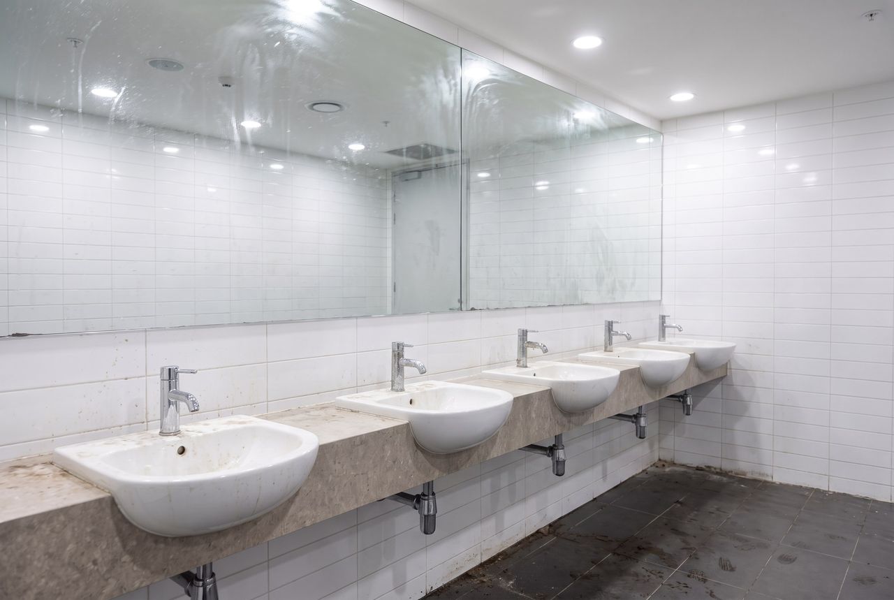 Commercial restroom overdue for a deep clean, limescaled chrome, a hazed mirror and darkened grout